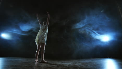 Elegant-ballerina-jumping-and-dancing-in-barefoot-and-white-loose-oversize-dress-in-studio.-With-smoke